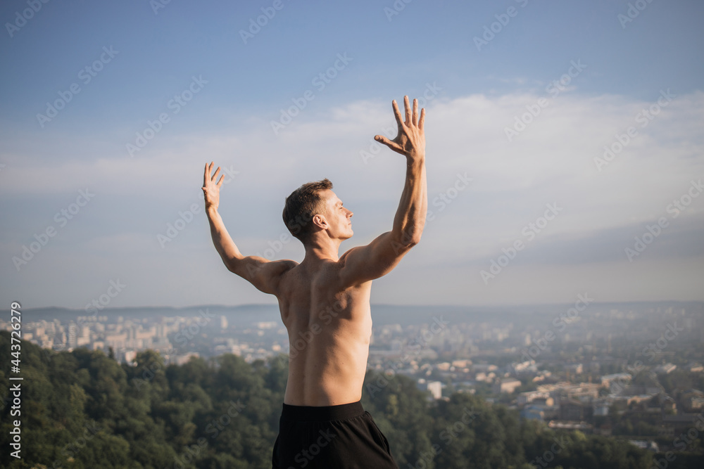 Strong young man with bare torso holding hands up while standing on high hill. Background of cityscape. Concept of active and healthy lifestyles. Strong young man with bare torso holding hands up while standing on high hill. Background of cityscape. Concept of active and healthy lifestyles.