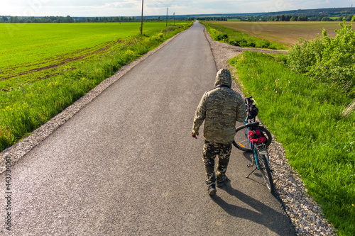 A cyclist in camouflage from the back next to a bicycle on the background of an asphalt road going into the distance