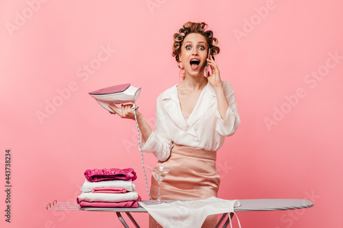 Lady talking on phone and opened her mouth in surprise. Housewife ironing clothes on pink background photo