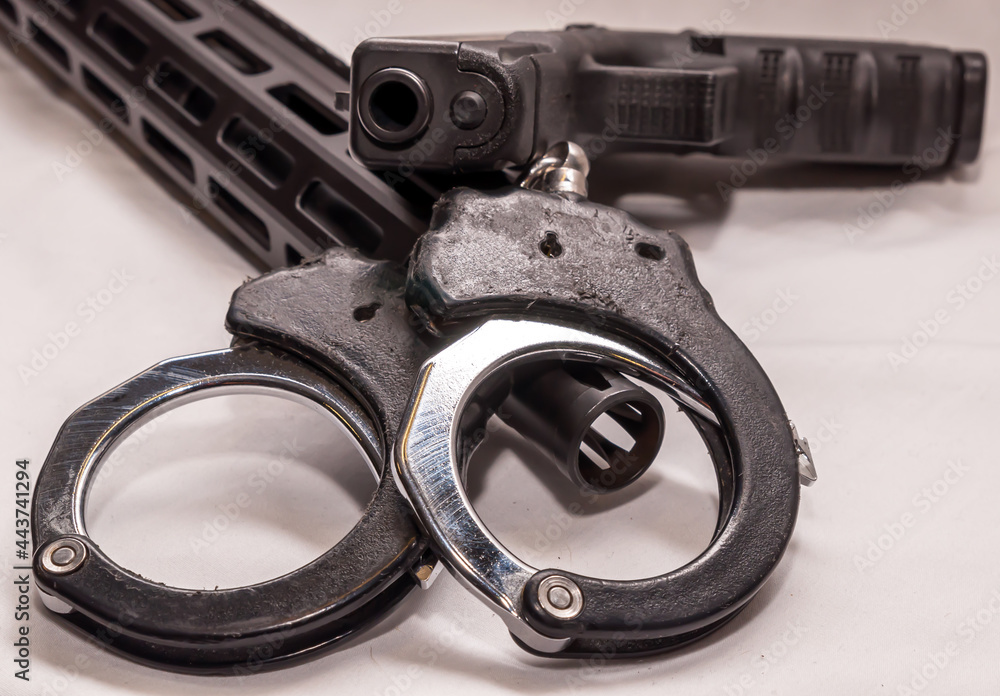 Obraz premium A pair of black and silver handcuffs laying over the barrel of an AR 15 rifle with a black 9mm pistol on top of them on a white background