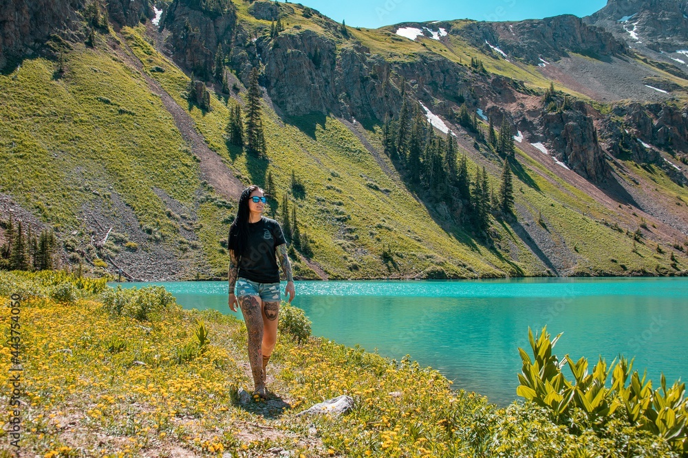 View of a woman with tattoos walking alongside the turquoise water of blue lake in Colorado. This is an alpine lake located along the Blue Lakes Trail.