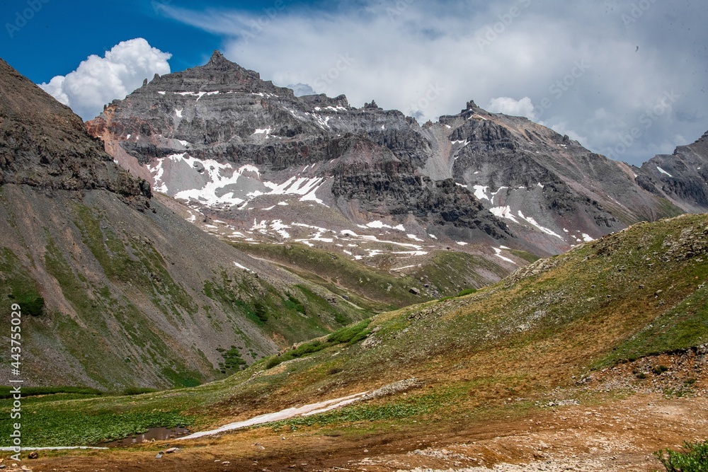 View of mountain peaks in Yankee Boy Basin near Ouray, Colorado. The mountain range has snow on it with jagged rocks, a blue cloudy sky, and green grass.