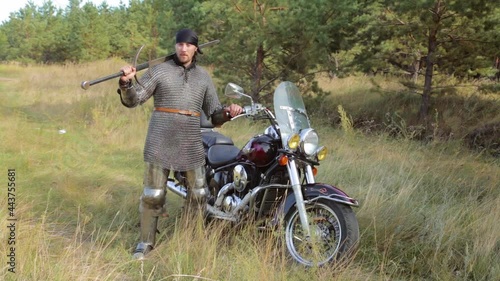 A medieval knight in a full set of armor and a bandana on his head poses with a two-handed sword in his hands while standing next to a motorcycle against the background of a forest.