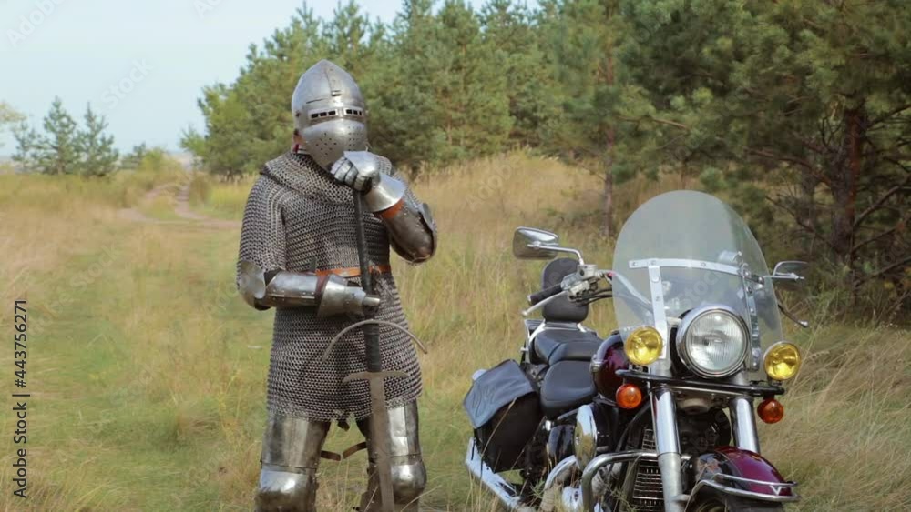 A medieval knight in full armor in chain mail with a helmet on his head and a two-handed sword in his hands poses next to a motorcycle against the backdrop of the forest. A medieval knight in full armor in chain mail with a helmet on his head and a two-handed sword in his hands poses next to a motorcycle against the backdrop of the forest.