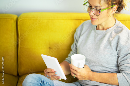 Close up portrait of mature pretty woman reading a book on tablet with glasses sitting on the yellow couch at home in break leisure time activity alone - adult people enjoy technology and learning