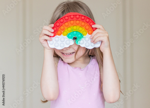 A little blonde girl holds a pop it toy in the shape of a rainbow in her hands and covers her face with it. Happy child peeking out from behind a toy