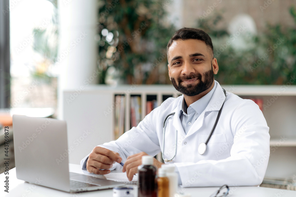 Portrait of an Indian general medicine doctor, sit at a desk in an office in a clinic, dressed in medical clothes with a stethoscope around his neck, looking at camera, smiles friendly. Medical expert Portrait of an Indian general medicine doctor, sit at a desk in an office in a clinic, dressed in medical clothes with a stethoscope around his neck, looking at camera, smiles friendly. Medical expert