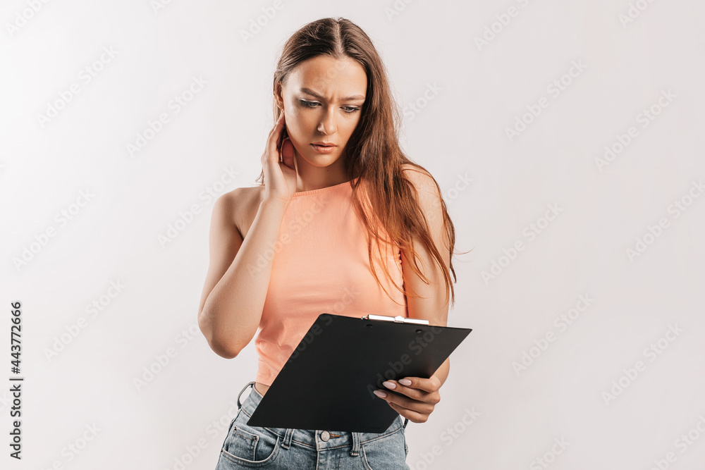 Girl student frowns with displeasure and reads information in documents. Beautiful young business woman holds a clipboard with mockup on gray background. Study and business concept. Online learning Girl student frowns with displeasure and reads information in documents. Beautiful young business woman holds a clipboard with mockup on gray background. Study and business concept. Online learning