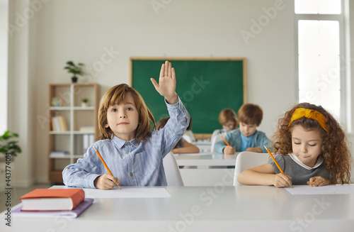 Boy and a girl are sitting at a school desk, writing, the boy raises his hand up. Primary school classmates are sitting in the classroom, the books are on the table, back to school. photo
