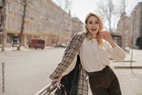 Excited blonde young woman in brown pants, white stylish blouse and checkered jacket leans on fence and looks surprised outside.