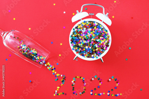 A glass with multicolored sequins, alarm clock and star shaped confettie on the red background. Party, new year, concept