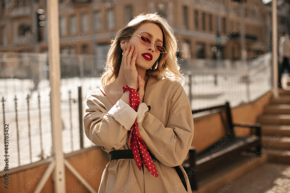 Fototapeta premium Gorgeous blonde elegant woman with red lips in bright colorful sunglasses and beige trench coat gently touches face and poses outside.