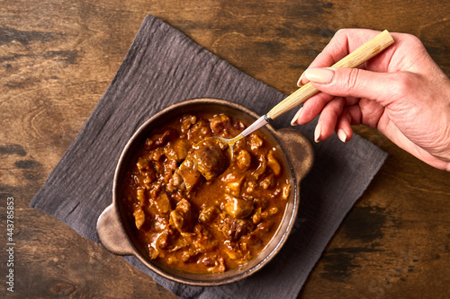 Woman's hand holds a fork with traditional goulash meat in ceramic bowl on napkin. Top view