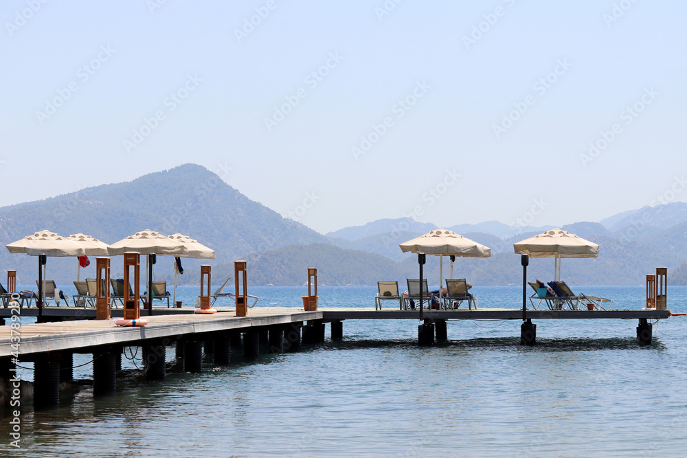 Wooden pier with lounge chairs and parasols on sea resort, vacation concept. People sunbathing on a beach on misty mountains background
