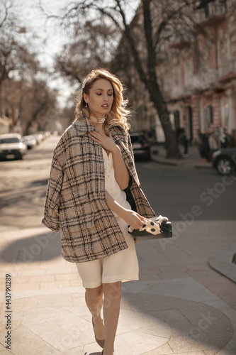 Young blonde curly woman in white dress, checkered tweed coat and pearl necklace walking in city and holding handbag.
