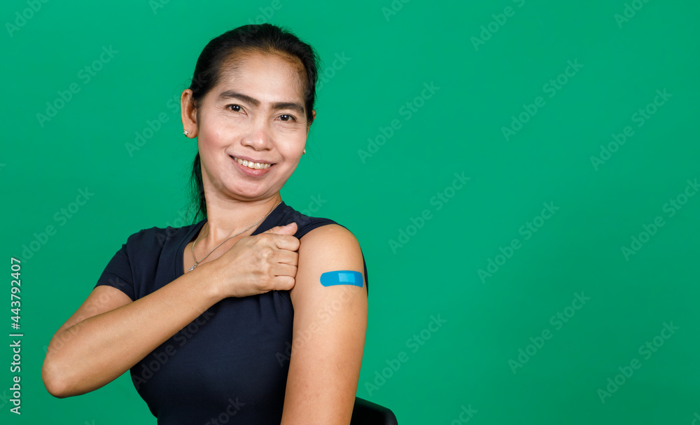 Middle aged Asian woman smiling showing her arm with bandage patch showing she got vaccinated for Covid 19 virus on green background. Concept for Covid 19 vaccination Middle aged Asian woman smiling showing her arm with bandage patch showing she got vaccinated for Covid 19 virus on green background. Concept for Covid 19 vaccination