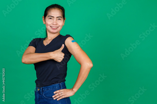 Asian middle aged woman smiling and pointing at her arm with bandage patch showing she got vaccinated for Covid 19 virus on green background. Concept for Covid 19 vaccination © Bangkok Click Studio