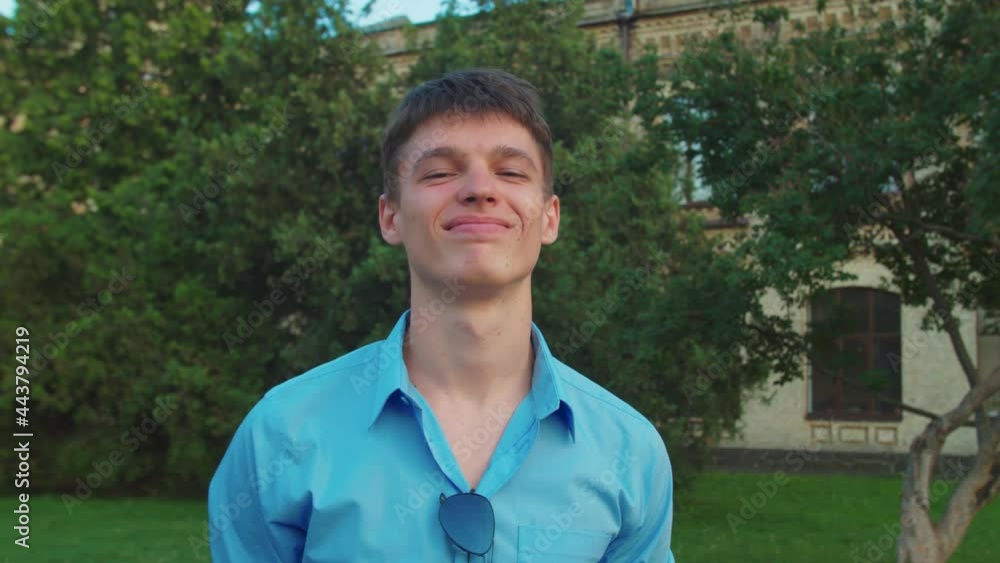 Close-up portrait of attractive young male student in casual clothing with forced smile looking directly into camera outdoors, having derisive look standing in front of old building at sunset Close-up portrait of attractive young male student in casual clothing with forced smile looking directly into camera outdoors, having derisive look standing in front of old building at sunset