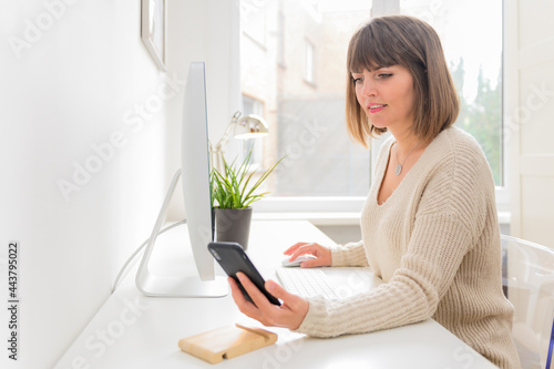 Charming young freelancer woman is talking on smartphone working with laptop from home, female employee has pleasant phone conversation working on the distance, connect with customers by mobile phone.