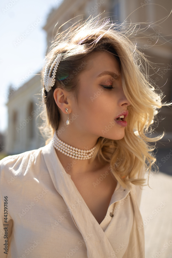 Fototapeta premium Young beautiful curly blonde woman in beige blouse and pearl necklace poses with closed eyes and walks outside.