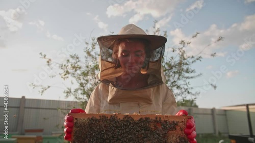 A girl beekeeper in a protective suit carries a frame with honey from a bee hive