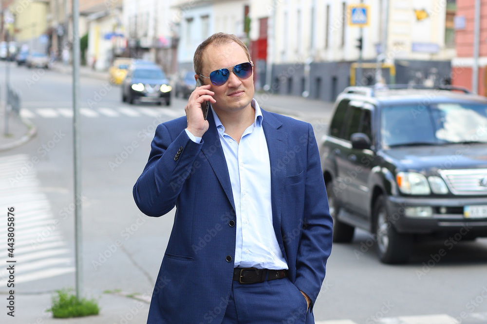 Banner with a young caucasian man in a blue business suit and sunglasses holds a smartphone for business negotiations. Work, business. Background - noisy center in the city.