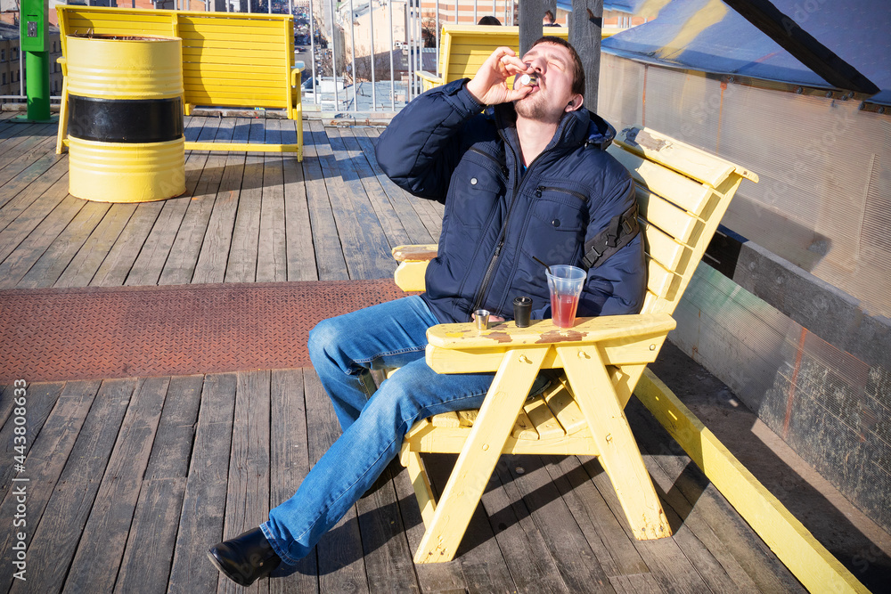 Young man drinking alcohol from little glass, head thrown back, sitting on wooden yellow armchair on sunny terrace in blue jeans and warm jacket. Young man drinking alcohol from little glass, head thrown back, sitting on wooden yellow armchair on sunny terrace in blue jeans and warm jacket.