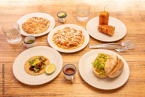 Plates of Mexican food, quesadillas, golden tacos, tacos al pastor, corn, cob, forks, sauces, sandwich.