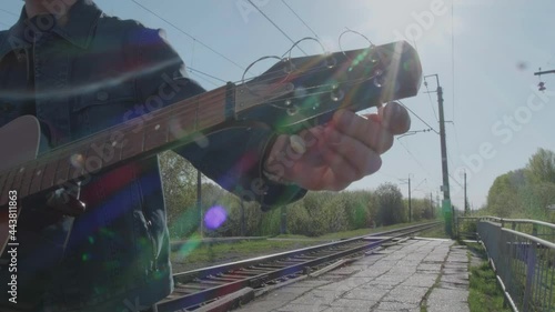 A man in a denim jacket tunes an acoustic guitar while standing next to the railroad tracks. Sun glare. light leak lens flare. Optical lens flare.