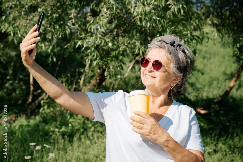 Fototapeta premium Modern smiling mature woman in red glasses and cup of coffee taking a selfie on mobile phone while standing in park, outdoors. Trendy beautiful senior woman using smartphone outside, summer lifestyle