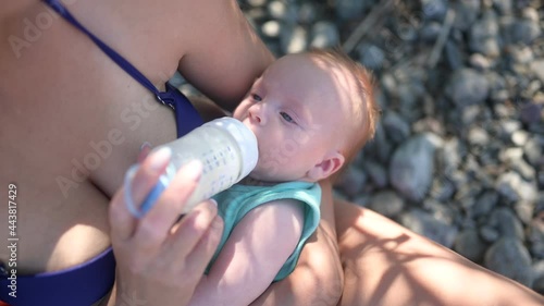 Young mother with newborn cute infant baby boy, holding him on arms, hugging and feeding with bottle breast milk outside in sunny day. Healthy child, concept of hospital and happy motherhood. Nursery.