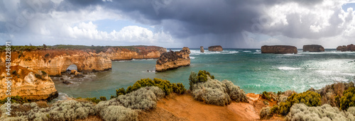 A huge panorama of rocky reddish cliffs and small islands in the southern ocean. Victoria. Great Ocean Road. A line of protruding rocky islands on the horizon. The sky is covered with dark clouds.
