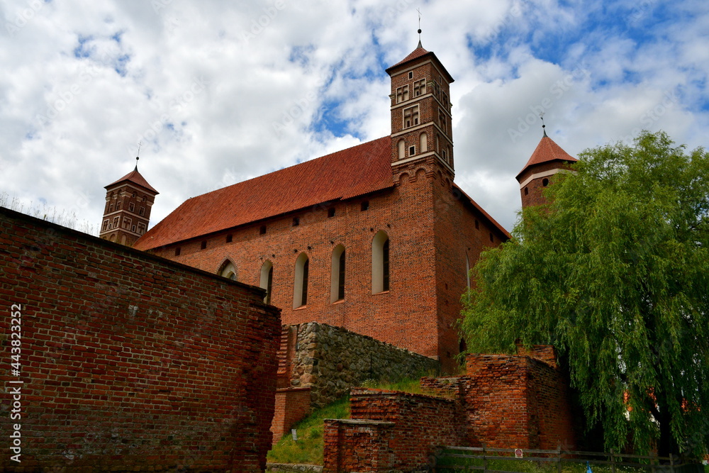 A close up on an old medieval castle made out red brick and stone with some towers and an angled roof seen next to the remnants of medieval walls that are partially damaged spotted in summer in Poland