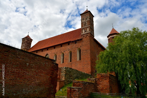 A close up on an old medieval castle made out red brick and stone with some towers and an angled roof seen next to the remnants of medieval walls that are partially damaged spotted in summer in Poland