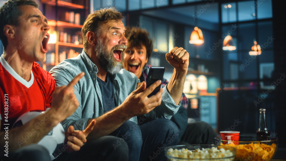 Night at Home: Three Soccer Fans Sitting on a Couch Watch Game on TV, Use Smartphone App to Online Bet, Celebrate Victory when Sports Team Wins. Friends Cheer Eat Snacks, Watch Football Play.