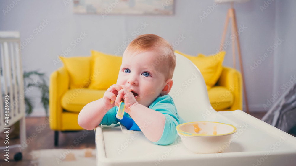 infant boy with blue eyes sitting in feeding chair and sucking spoon near bowl infant boy with blue eyes sitting in feeding chair and sucking spoon near bowl