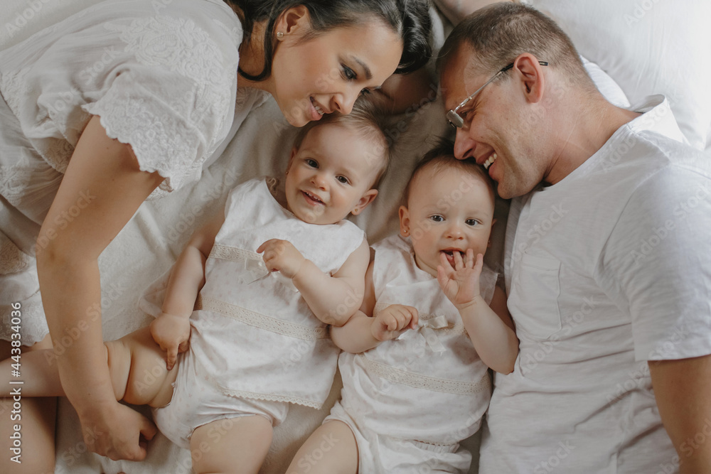 Top view of charming family portrait with baby twins. Smiled parents with firsborns in love dressed in white clothing, laying at bedroom, weekend, family vacation. Concept of happy healthy parenthood