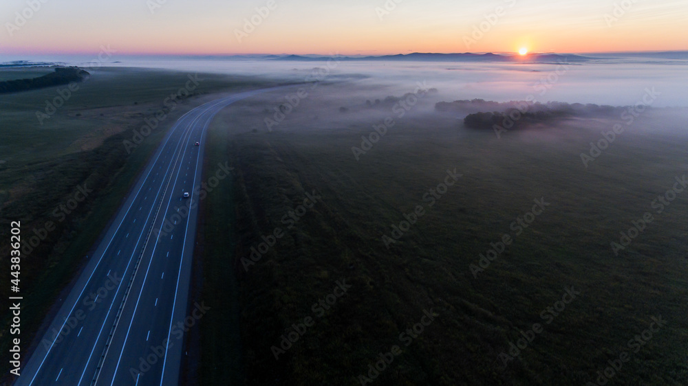 View from above. An asphalt road is located among endless green meadows. Automobile road in the fog against the background of dawn.