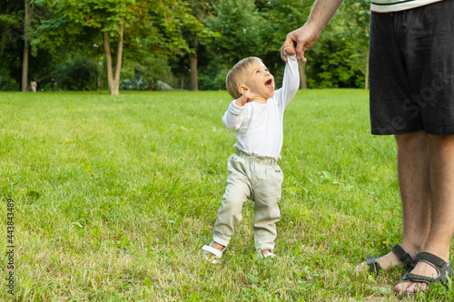 Happy caucasian blond baby girl standing on grass looking at unreconizable man, laughing, smiling. Dad and kid walking in park. Father and child, spending time together in summer. copy space for text
