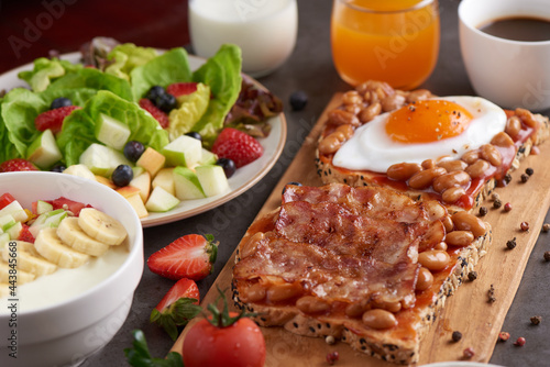 variety of open sandwiches made of brown whole wheat  bread with tomatoes sauce, bowl of oat granola with yogurt, white beans, bacon, fried egg. fresh fruit and vegetable salad, healthy breakfast.
