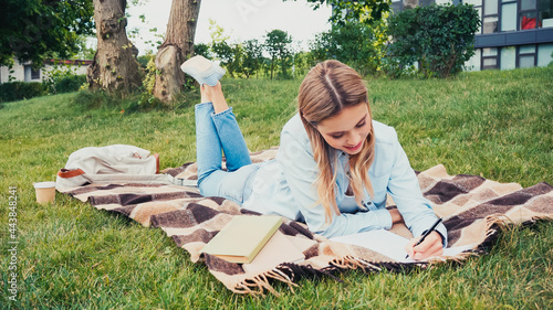 cheerful student writing in notebook near books while lying on blanket in park