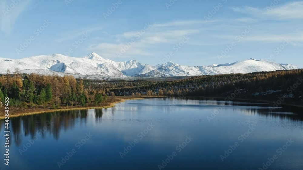 Blue lake, autumn forest and snowy mountains on horizon at sunny day. Flying over quiet pond among woods, cloudy sky and white peaks are reflected in serene mirror surface of water Blue lake, autumn forest and snowy mountains on horizon at sunny day. Flying over quiet pond among woods, cloudy sky and white peaks are reflected in serene mirror surface of water