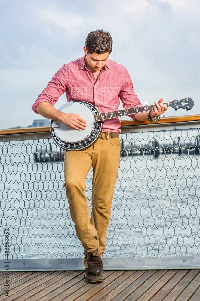 Dressing in light red shirt, dark yellow trousers and brown shoes, hanging his head and stepping his foot, a young musician is playing a banjo on the deck