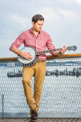 Dressing in light red shirt, dark yellow trousers and brown shoes, looking down and stepping his foot, a young musician is playing a banjo on the deck