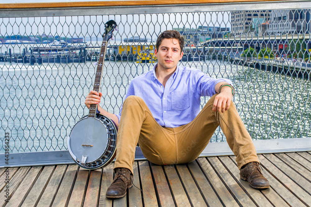 Dressing in a light blue shirt, dark yellow jeans and brown boot shoes, holding a banjo, a young musician is sitting on the deck against a fence, relaxing