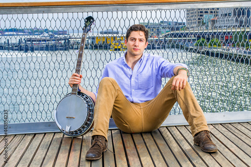 Obraz na płótnie Dressing in a light blue shirt, dark yellow jeans and brown boot shoes, holding a banjo, a young musician is sitting on the deck against a fence, relaxing w salonie