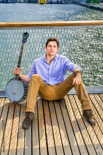 Dressing in a light blue shirt, dark yellow jeans and brown boot shoes, holding a banjo, a young musician is sitting on the deck against a fence, relaxing