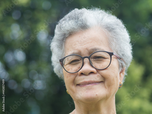 Portrait of an elderly Asian woman with short gray hair, wearing glasses, smiling and looking at the camera while standing in a garden. Aged people and relaxation concept