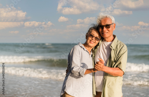 Asian senior couple tourist is hand in hands, embrace and dancing on tropical sea beach in summer holiday.