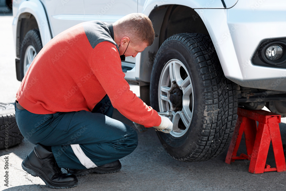 A car mechanic in a medical mask changes a wheel on a car on the street on a sunny day. Help on the road with a tire puncture. Urgent tire service A car mechanic in a medical mask changes a wheel on a car on the street on a sunny day. Help on the road with a tire puncture. Urgent tire service
